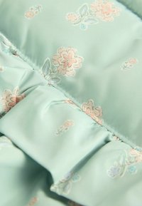 Helles mintgrünes Gewebe mit einem Blumenmuster, das rosa und blaue Blumen zeigt. Enthält geraffte Details und eine glatte Textur.