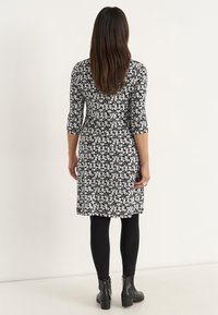 Kvinna med långt brunt hår, klädd i en svart och vit blommig klänning, svarta leggings och svarta ankelboots, stående vänd mot en ljus vägg.