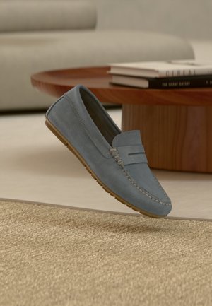Mocassin en daim bleu avec détails cousus, présenté près d'une table basse en bois et d'un tapis beige dans un salon moderne.