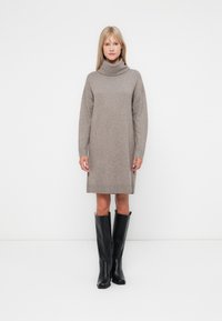 Vestido de malha em castanho claro, com gola alta, mangas longas e punhos canelados. Combinado com botas de couro pretas até ao joelho.