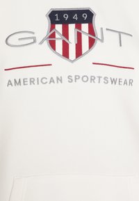 Baltas gobtuvas su siuvinėtu GANT logotipu, vaizduojančiu šalmą tamsiai mėlynos ir raudonos spalvos, kartu su tekstu "AMERICAN SPORTSWEAR" pilka spalva.