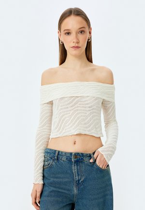 Crop top na ramiączkach wykonany z białej fakturowanej dzianiny, z długimi rękawami i falistymi wzorami, w zestawie z niebieskimi dżinsami z wysokim stanem.