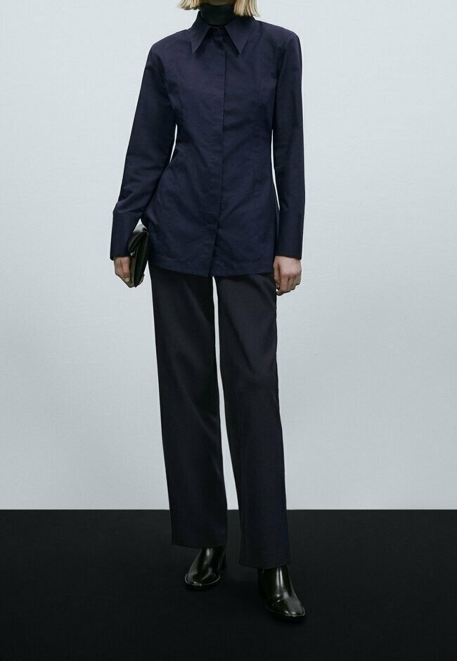 Personne portant une chemise bleu marine foncé à manches longues avec col, pantalon large assorti, bottines noires, tenant une pochette noire.