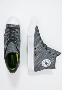 Vysoké tenisky v černé a šedé texturované látce s bílými gumovými podešvemi a kroužky. Obsahují kulatou nášivku loga na vnější straně.