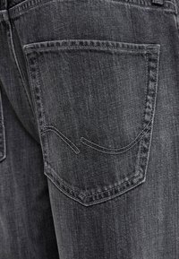 Tumši pelēkas džinsa bikses ar vienu aizmugurējo kabatu, kurai ir izliekti šuvju rotājumi un maigs teksturēts apdarinājums.