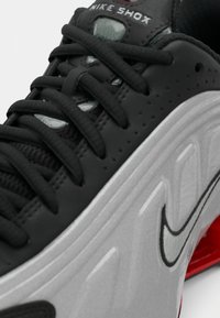 Close-up de um ténis Nike Shox preto e cinza com atacadores pretos e amortecimento vermelho visível na sola.