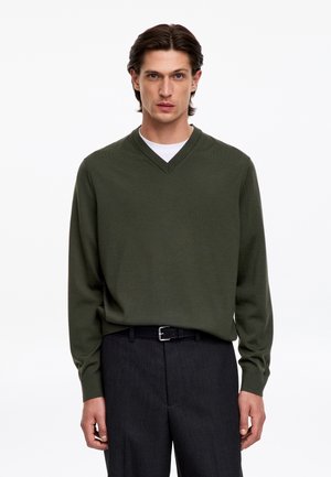 Homme aux cheveux foncés portant un pull vert foncé à col en V par-dessus une chemise blanche et un pantalon foncé avec ceinture noire, debout devant un fond blanc uni.
