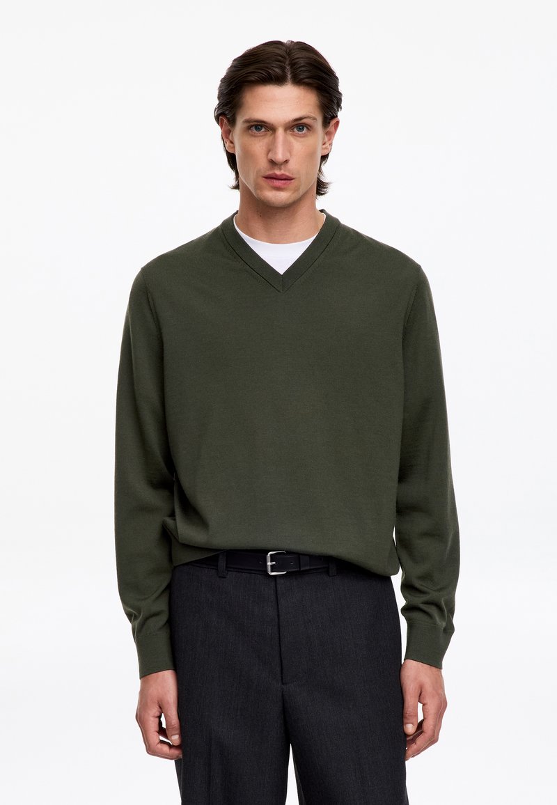 Homme aux cheveux foncés portant un pull vert foncé à col en V par-dessus une chemise blanche et un pantalon foncé avec ceinture noire, debout devant un fond blanc uni.