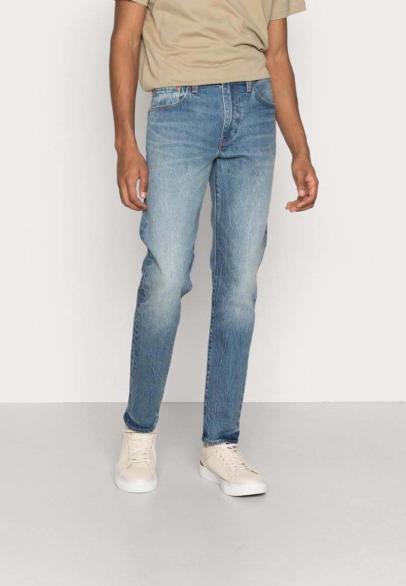 Ljust blå denimjeans med slim fit, featuring subtila blekningar och slitningar, kombinerat med en beige t-shirt och vita sneakers.
