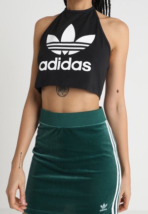 Czarny bawełniany krótki top z wiązaniem na szyi z białym logo Adidas i wzorem trzech pasków. Do tego zielona aksamitna spódnica z białymi detalami.