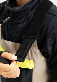 Salopette imperméable avec un devant beige, une section inférieure noire et une poche jaune vif. Équipée de bretelles noires ajustables et d'un tissu texturé.
