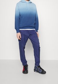Sudadera con degradado azul y un bolsillo frontal, combinada con pantalones cargo azul marino y zapatillas negras con grandes letras blancas y detalles en rojo.