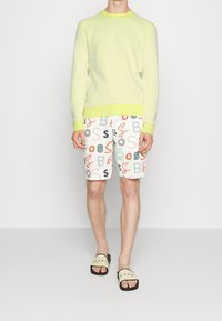 Jasnozielony dzianinowy sweter z okrągłym dekoltem, zestawiony z białymi szortami z kolorowymi logo "BOSS" oraz czarnymi klapkami z czerwonym napisem.