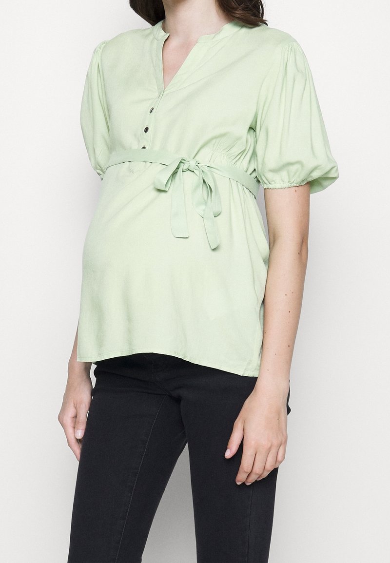Blouse vert clair avec un col en V, des manches courtes bouffantes, des boutons sur le devant et une ceinture à nouer à la taille. Associée à un pantalon noir ajusté.