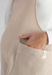 Lange beige schort gemaakt van een geweven wafelstof. Heeft een voorzak en een label, met een witte lange mouwtop die gedeeltelijk zichtbaar is eronder.