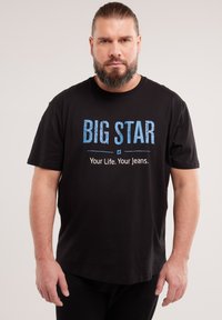 Uomo barbuto con capelli lunghi raccolti indietro che indossa una maglietta nera con la scritta "BIG STAR Your Life. Your Jeans." su uno sfondo chiaro uniforme.