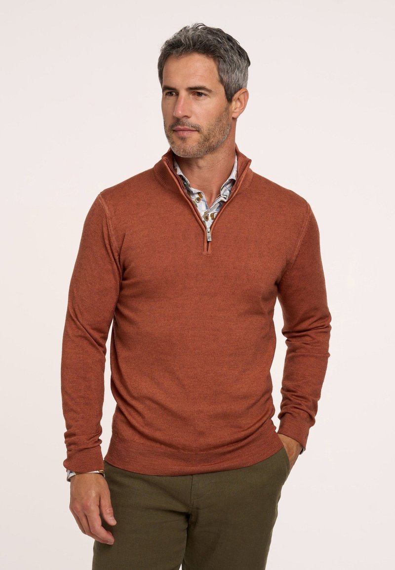 Pullover in Rostfarbe mit Reißverschluss am Hals, aus weichem Strickstoff gefertigt. Verfügt über lange Ärmel und eine figurbetonte Passform, kombiniert mit einem gemusterten Hemd darunter.