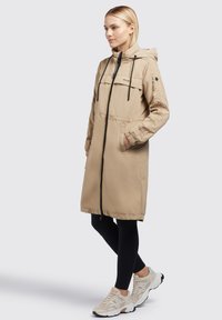 Beige Kapuzenparka mit einem Reißverschluss vorne, elastischen Bündchen und zwei Seitentaschen. Mit schwarzen Kordeln und einem Logo-Akzent auf der Brust.