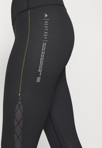 Svarta atletiska leggings med en slät yta, som har gula sömmar, nätutskärningar och tryckt text som anger "HEAT.RDY" teknologi.