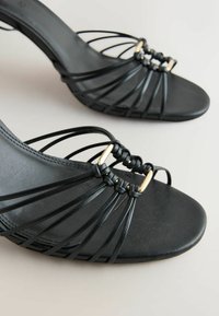 Sandales à talons hauts noires à bout ouvert avec fines lanières et boucle rectangulaire dorée sur fond clair.