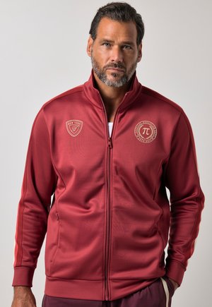 Mann mit kurzen Haaren und Bart, trägt eine rote Sportjacke mit Reißverschluss, bedruckt mit Logos auf der Brust und Streifen an den Ärmeln vor einfarbigem Hintergrund.