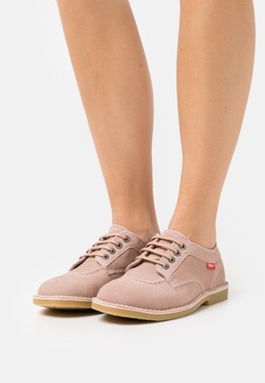 Chaussures en daim rose clair avec fermeture à lacets, semelle en caoutchouc, et une petite étiquette rouge. Bout rond avec un détail de couture et une texture lisse.