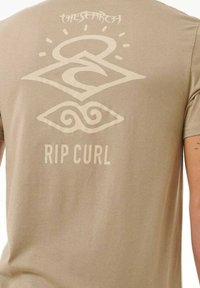 Maglietta in cotone beige con una grande grafica chiara sul retro, comprensiva del testo "RIP CURL" e design astratti.