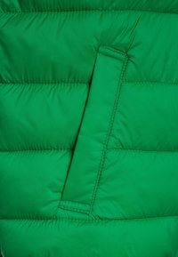 Tessuto verde trapuntato con cuciture orizzontali e una tasca rettangolare verticale con dettagli di cucitura visibili.