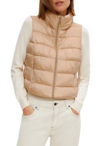 Béžová prošívaná vesta s vysokým límcem, vybavená předním zipem a bočními kapsami. Nositelná přes bílý dlouhý rukáv.