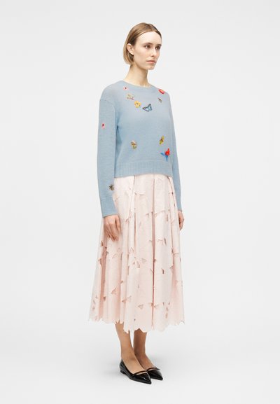 Γυναίκα που φοράει ένα ανοιχτό μπλε πουλόβερ με κεντήματα πεταλούδων και μια ανοιχτό ροζ φούστα με κοψίματα, σε συνδυασμό με μαύρες μυτερές μπαλαρίνες.