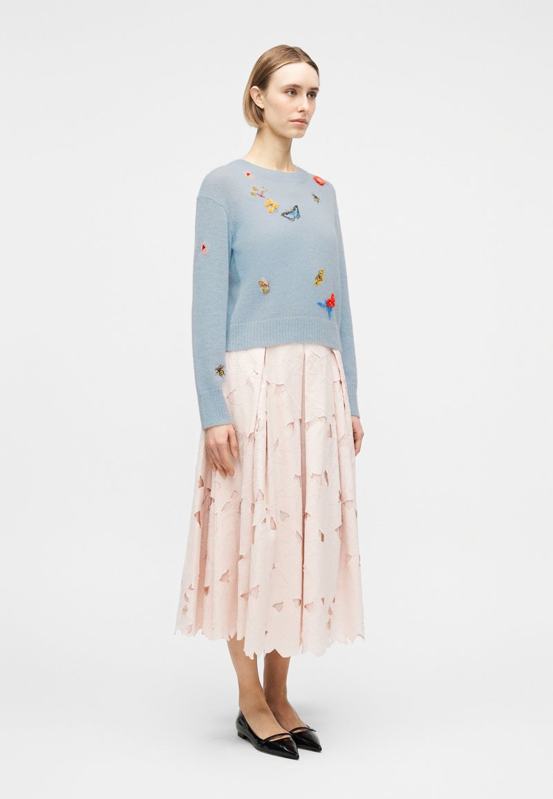 Donna che indossa un maglione azzurro chiaro con ricami di farfalle e una gonna rosa pallido con motivi traforati, abbinati a ballerine nere con punta a stivaletto.