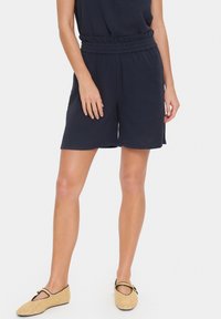 Marineblaue Shorts mit strukturiertem Finish, elastischem Bund und seitlichen Schlitzen. Kombiniert mit hellbraunen, geflochtenen Schuhen mit einer Schleifendetail.