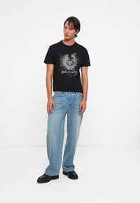 Črna majica s kratkimi rokavi z sivo grafično logotipom spredaj, v kombinaciji s svetlo modrimi širokimi kavbojkami in črnimi usnjenimi čevlji.