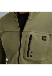 Zeleno zip-up jopico z rebrastim ovratnikom, teksturirana tkanina, prsni žep z zadrgo in subtilna oznaka blagovne znamke na žepu.