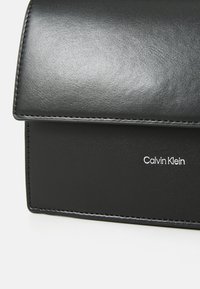 Sac à main en cuir noir avec une texture lisse, de forme rectangulaire et un rabat pliable. Présente des coutures subtiles et un logo "Calvin Klein" blanc.