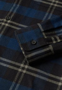 Camicia di flanella a quadri con un motivo blu scuro, navy e grigio. Tasca frontale con bottone, texture liscia e polsini ripiegabili.