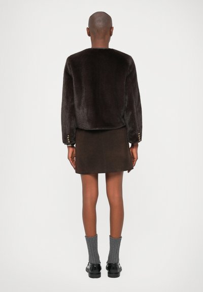 Brun kunstpelsjakke med lange ermer, avrundet kant og knappdetaljer på mansjettene, kombinert med en brun mockaskinn miniskjørt og grå ribbestrikkede sokker.