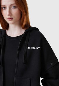 Sudadera negra con cordón, con cierre de cremallera y bolsillos frontales. "ALLSAINTS" impreso en blanco en la parte superior izquierda. Textura suave.
