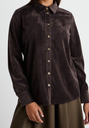Vrouw die een donkerbruine corduroy blouse met knopen draagt, met lange mouwen en een kraag, gecombineerd met een olijfgroene rok.