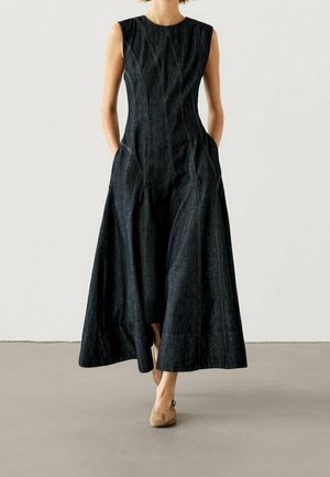 Kobieta w bezrękawną, ciemnoniebieską sukienkę midi w kształcie litery A z widocznymi szwami, nosząca beżowe baleriny, stoi na tle jednolitego tła.