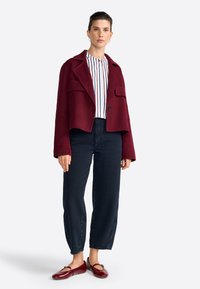 Veste bordeaux courte avec de grandes poches, assortie à un blouse rayée et un jean taille haute bleu foncé. Des chaussures rouges brillantes complètent le look.