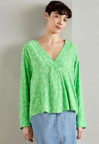 Grønn langermet topp med V-hals, med et hvitt blomstermønster. Stoffet ser glat og lett ut, med en avslappet passform.