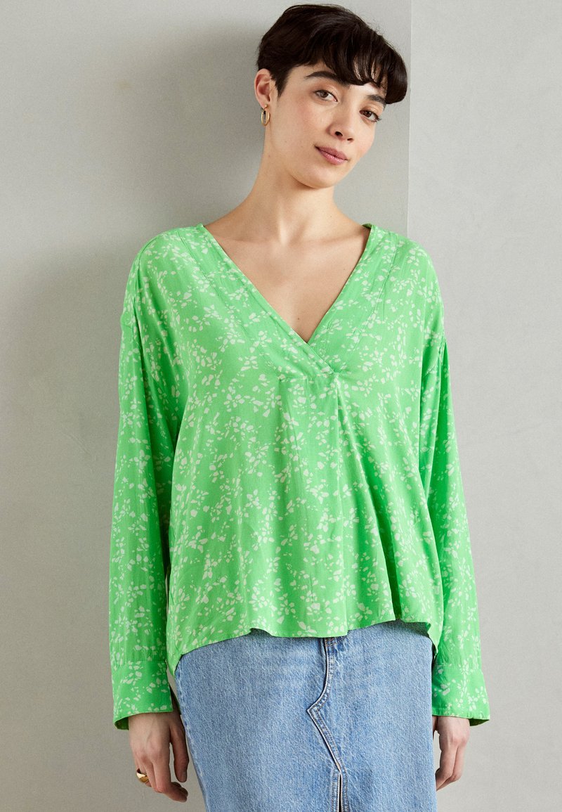 Grønn langermet topp med V-hals, med et hvitt blomstermønster. Stoffet ser glat og lett ut, med en avslappet passform.