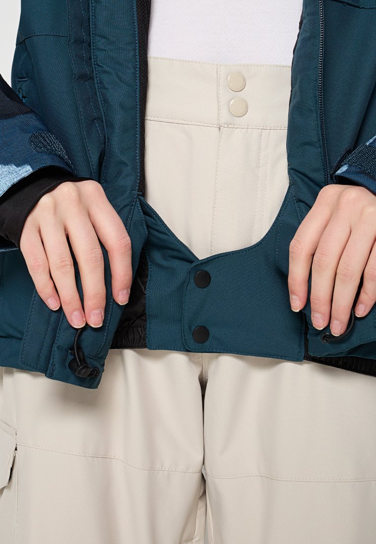 Türkise isolierte Jacke mit schwarzen Akzenten, ausgestattet mit Druckknöpfen und einem verstellbaren Saum, die über beigen Hosen mit strukturierter Oberfläche getragen wird.