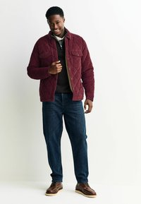 Bordeauxkleurige gewatteerde jas met twee voorke pockets, gedragen over een donkere trui en gecombineerd met blauwe jeans en bruine veterschoenen.