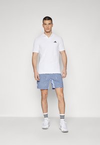 Camisa polo blanca con un logo negro, combinada con pantalones cortos azules estampados con un diseño ondulado. Completado con zapatos blancos y calcetines a rayas.