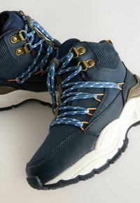 Sneakers înălțați de culoare bleumarin, cu material texturat, șireturi cu un model albastru și alb, accente de metal aurit și o talpă albă robustă.