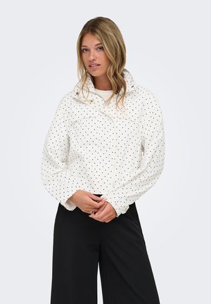Jonge vrouw met lang blond haar, een witte jas met stippen en een zwarte broek, staand tegen een effen achtergrond.