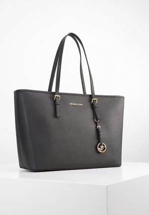 Czarna torba typu tote Michael Kors z dwoma paskami na ramię, złotym emblematem logo i wiszącym okrągłym ozdobnym elementem MK, prezentowana na białej powierzchni.