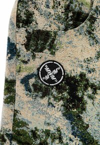 Tejido texturizado con un patrón inspirado en la naturaleza en verde y azul sobre un fondo beige, con un destacado parche de logo circular negro.